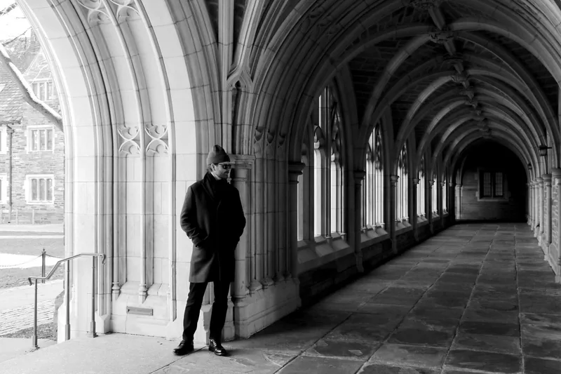 A man in a long dark coat leaning against a stone pillar in a Gothic cloister, arches receding into the distance behind him.