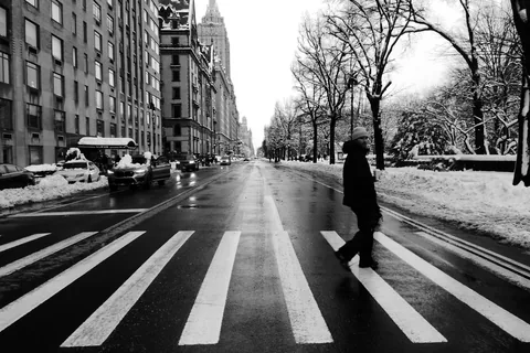 A lone figure crossing a wide Manhattan avenue on a snowy day, crosswalk stripes running toward the camera.