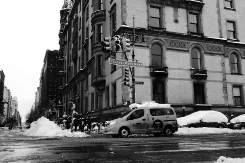 A snowy Manhattan street corner with a taxi van parked beside a stone apartment building and a traffic light overhead.