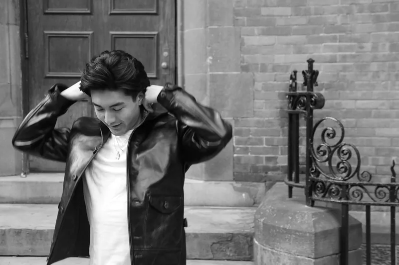Portrait of a young man in a leather jacket outside a stone building, hands raised behind his head, looking down.